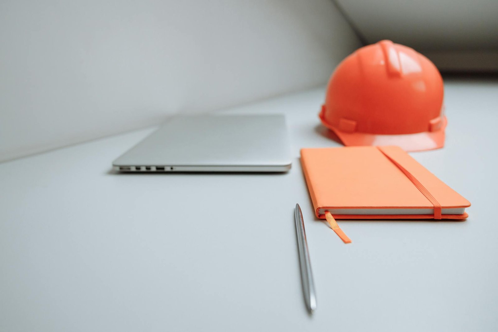 A minimalist office setup featuring a laptop, orange notebook, and safety hard hat.